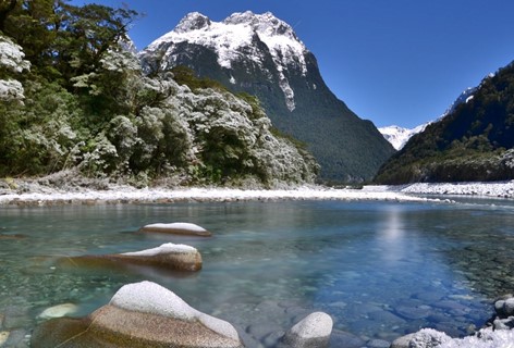 The clear blue river beside Milford Sound Lodge with snow settles on the tree, rocks and peaks.