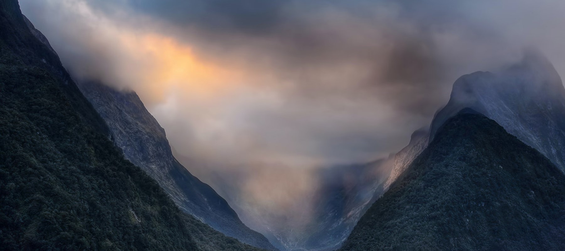 The sun breaks through grey clouds over Milford Sound between mountain peaks.