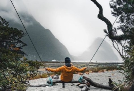 A child enjoys a swing on the banks for Milford Sound with mountain peaks in the background.