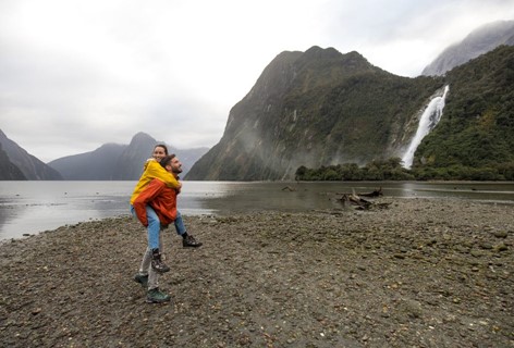 A couple walks along the stony Milford Sound shore with a waterfall in the background.