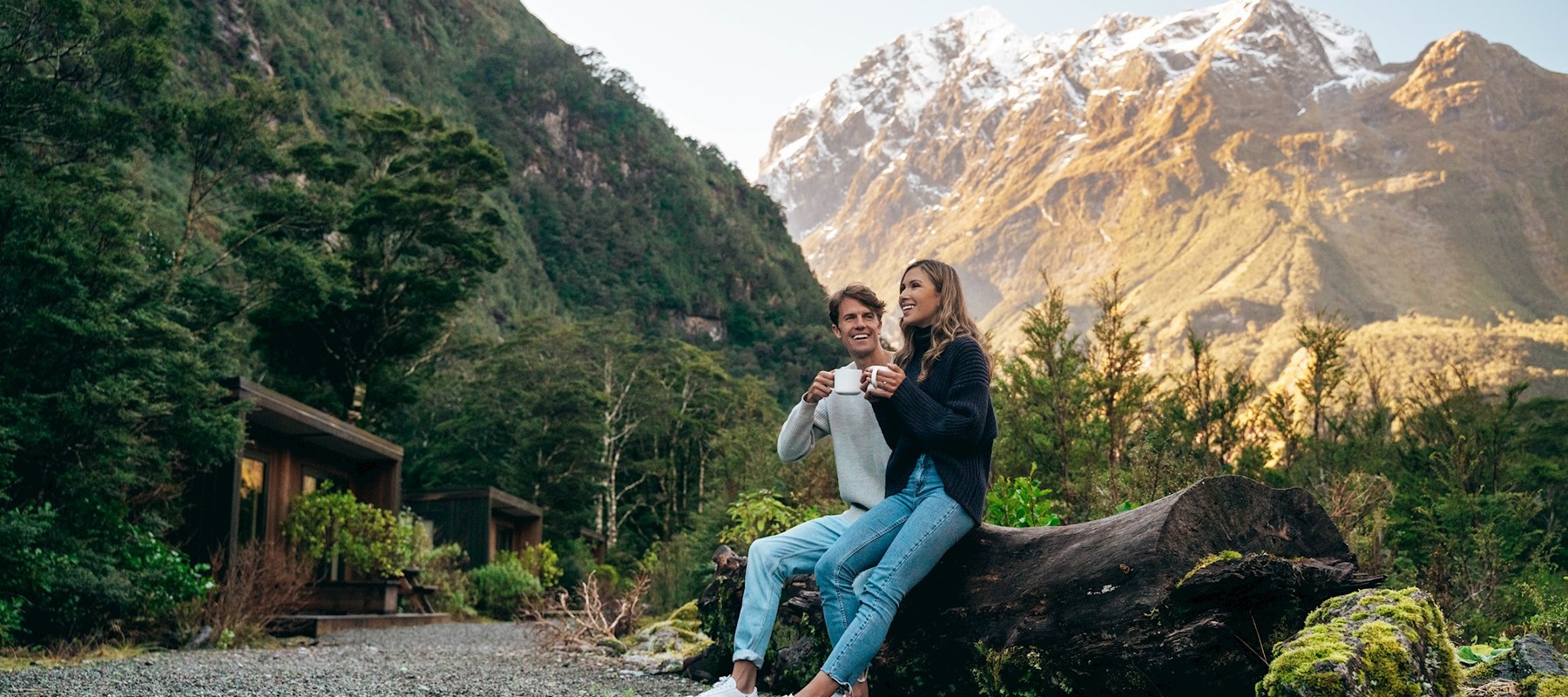 A couple enjoying a hot coffee sitting on a log outside the Milford Sound Lodge Mountain View Chalets 