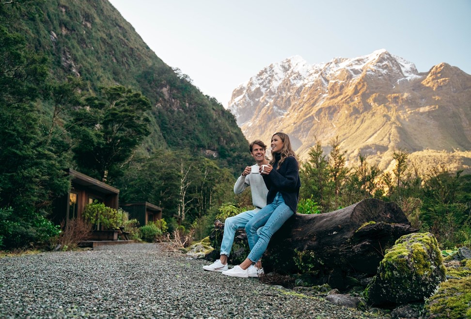 A couple enjoying a hot coffee sitting on a log outside the Milford Sound Lodge Mountain View Chalets 