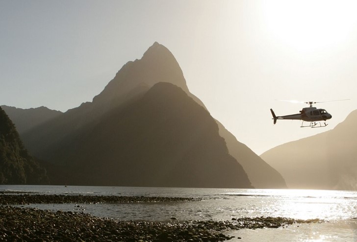 A helicopter flies above the Milford Sound near sunset between mountain peaks.