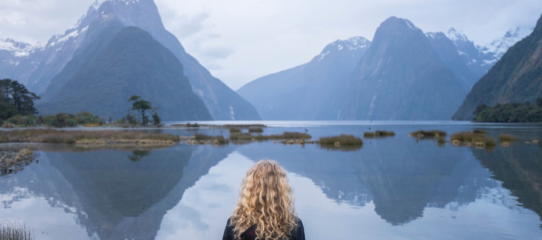 A curly-haired female looks across the Milford Sound peaks and calm water.